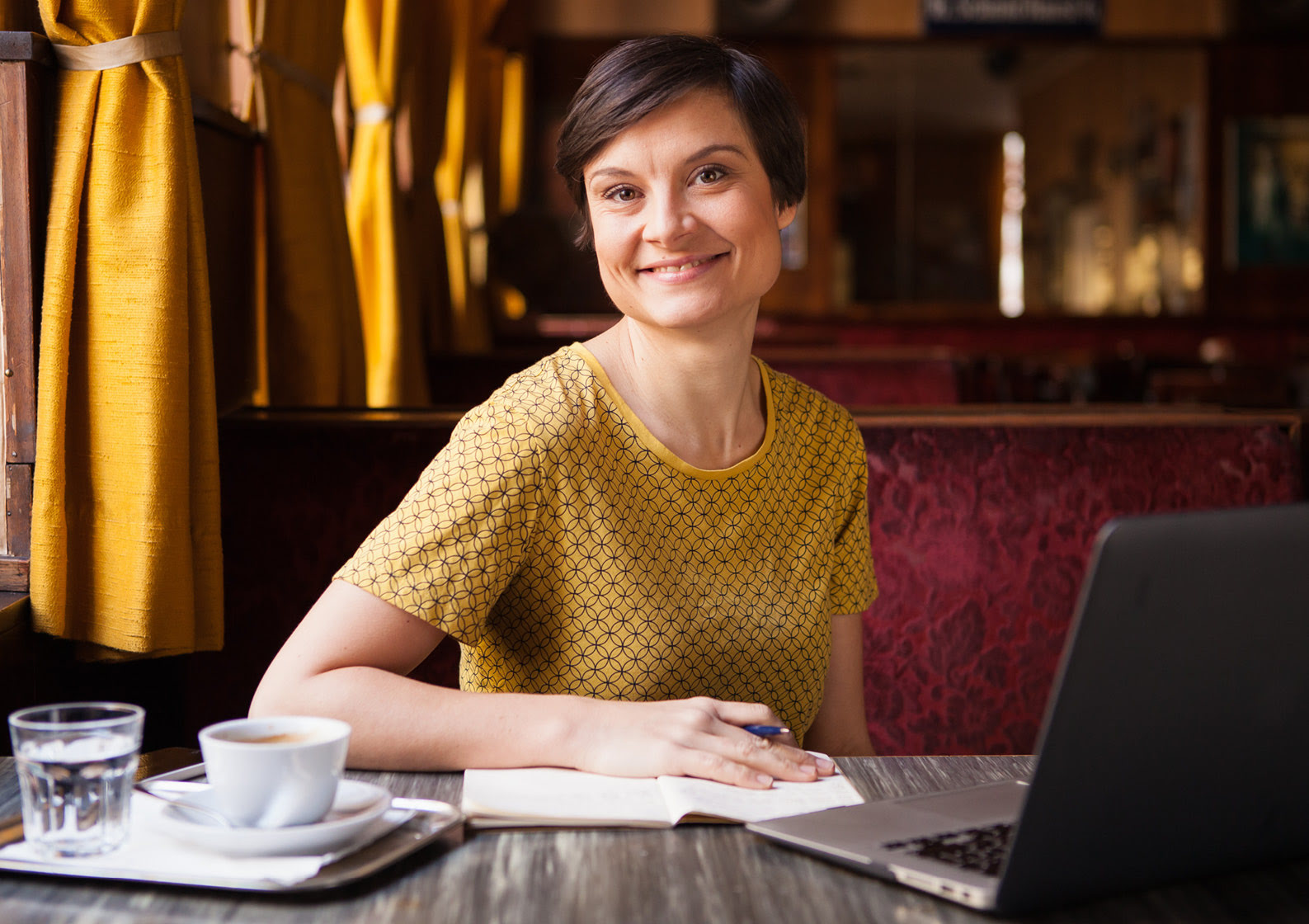Lena Raubaum sitzt mit Kaffee, Wasserglas, Notizbuch und Laptop entspannt in einem elegant eingerichteten Café mit samtenen, roten Bänken und senfgelben Vorhängen.