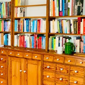 Bücherregal Heinrich beherbergt viele bunte Bücher auf seinen hölzernen Regalbrettern. Eine Kaffekanne und ein Heferl stehen auf seinem Unterbau.
